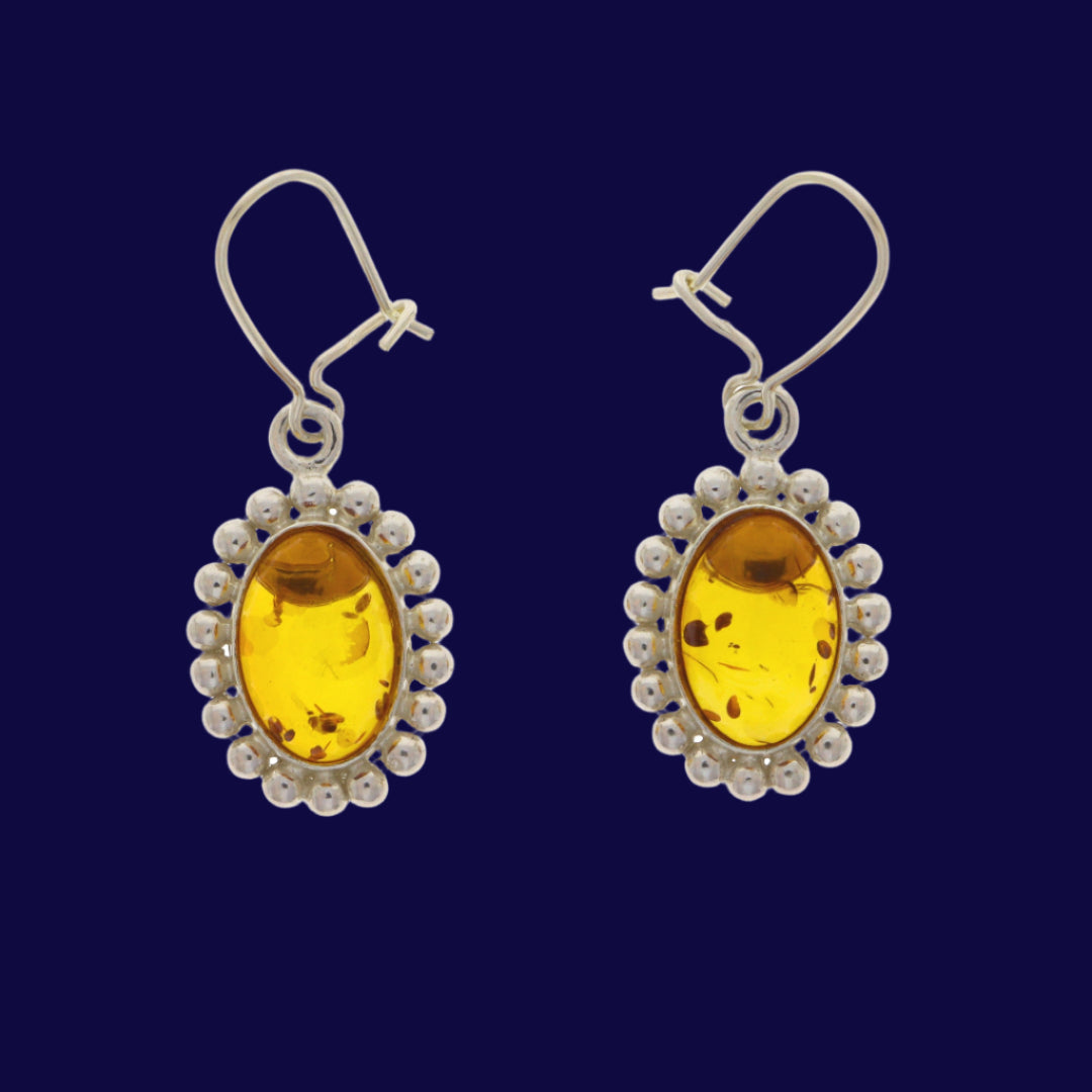 These earrings feature an oval, translucent yellow amber cabochon, surrounded by a distinct border of small, uniform silver spheres or beads, resembling a beaded edge or milgrain work, but more pronounced.
The overall design, with its oval shape and the beaded halo effect, gives them a classic, almost Victorian or Edwardian revival feel, reminiscent of traditional cameo settings or framed miniatures, yet simplified for a timeless appeal.
